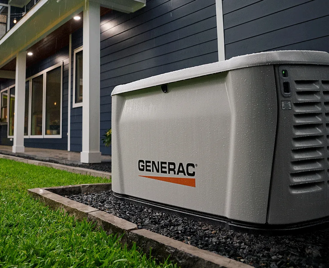 Generac home standby generator installed on a gravel pad beside a two-story home with a lit back porch